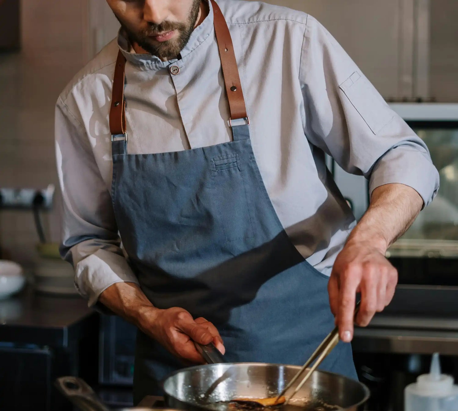 Chef portant un tablier bleu-gris avec des bretelles en cuir en train de cuisiner.